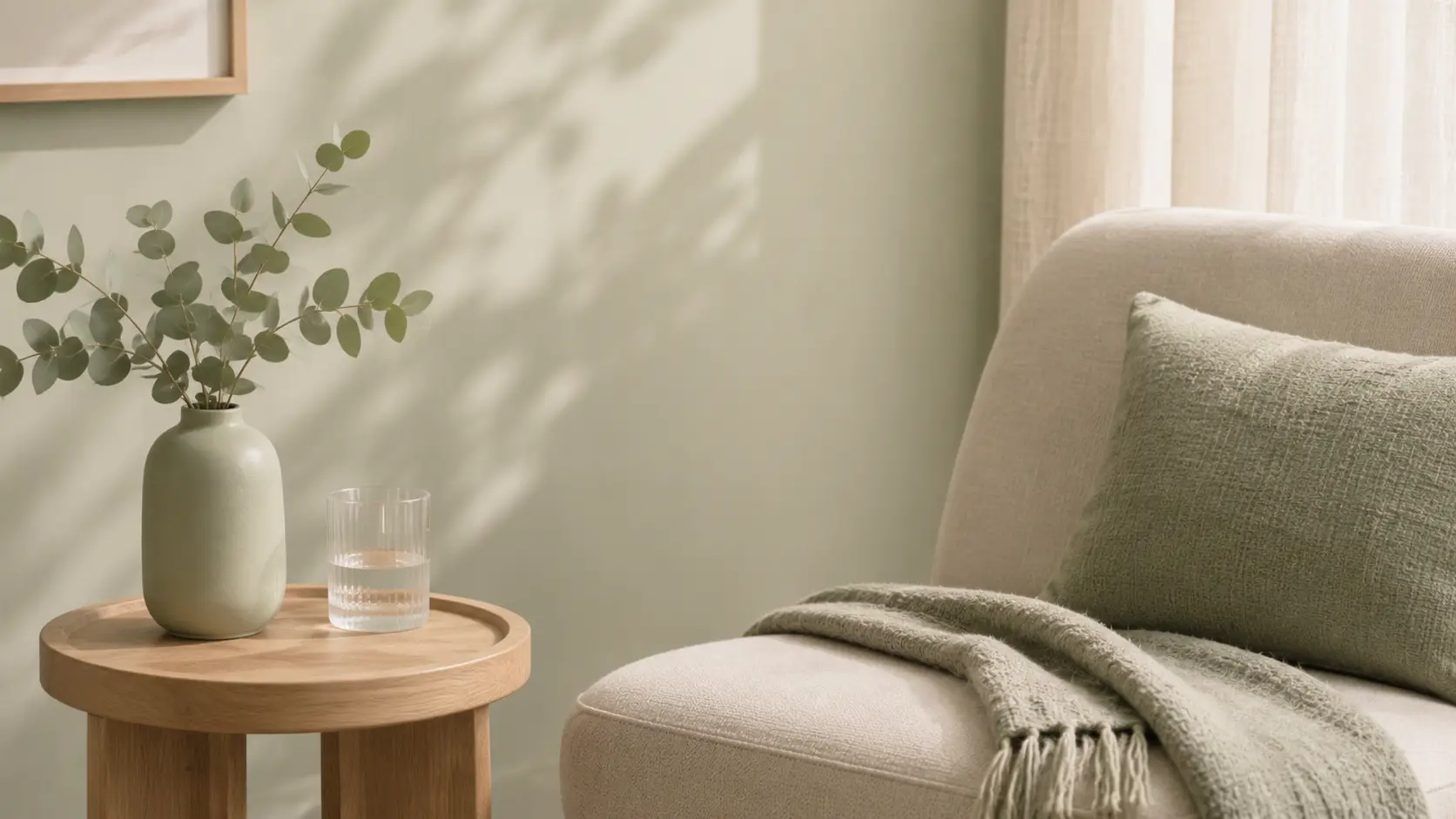 A sunlit corner of the room: eucalyptus in a pale green vase and a glass of water on a round oak side table, beside an armchair with a sage cushion and a soft throw.